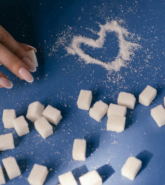 Top view of sugar cubes and a heart drawn in sugar on a blue surface with a hand nearby.